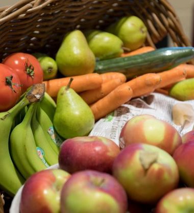 A variety of fruits and vegetables are spilling out of wooden basket onto a red table.