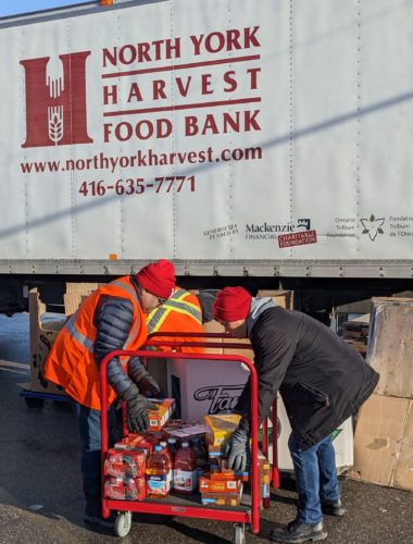 Two men dressed in winter clothes are moving food onto a trolley. A truck with North York Harvest Food Bank is in the background.