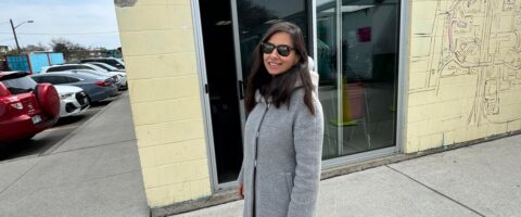 A woman with long dark hair wears sunglasses and smiles outside the Lawrence Heights food space.
