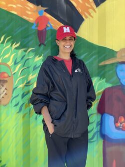 A woman wearing a red North York Harvest hat stands smiling outside in front of a brightly painted shipping container