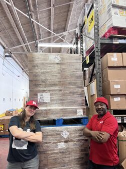 A man and woman wearing red North York Harvest hats smile with arms crossed in front of a large stack of menstrual products in a warehouse.