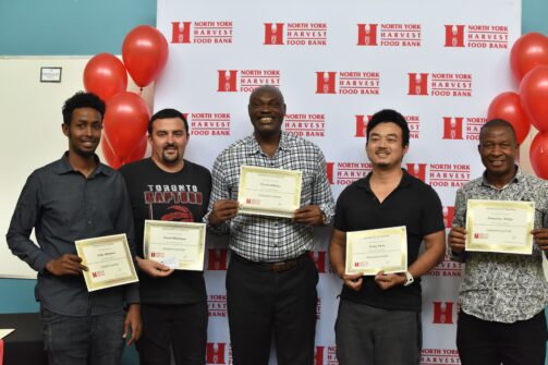 Five people holding graduation certificates stand in front of a back drop with the North York Harvest Food Bank logo on it. Red balloons are visible behind them. They are standing in a line, smiling for a picture.