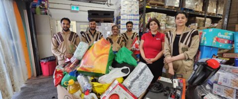 A group of individuals wearing Green S uniforms stand in front of a large bin of donated food, smiling for the camera with a representative from North York Harvest Food Bank.