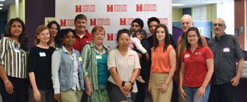 A group of people stand in front of a North York Harvest Food Bank backdrop, smiling for the camera.