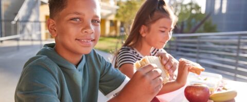 Two children sit outside eating lunch. A boy holding a sandwich is in the foreground, smiling.