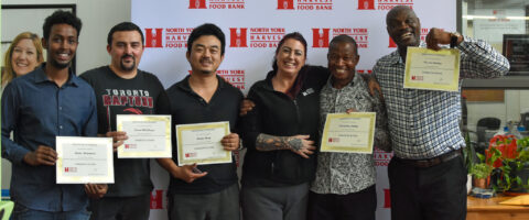 Six people stand in front of the North York Harvest Food Bank backdrop, five of whom are holding certificates.