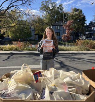 A teenage girl stands outside on a sunny day in front of a box full of bags of donated food. She holds a poster for her food drive which reads 'We Scare Hunger'