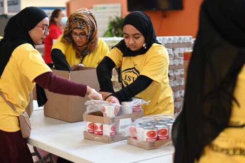 A group of volunteers wearing yellow Islamic Society of Vaughan t-shirts pick up cans and place them in boxes.