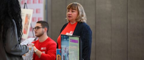 A person in a red shirt stands behind an information booth speaking to a person.