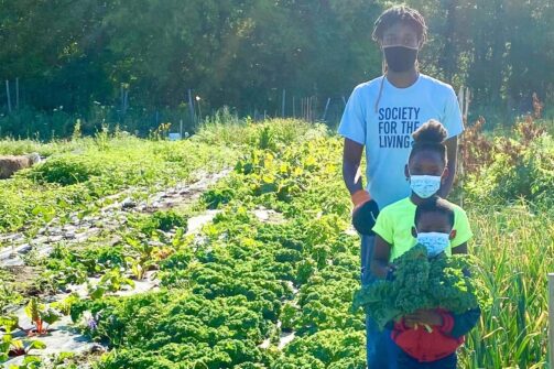 A father and daughter stand outside at a farm holding fresh vegetables.