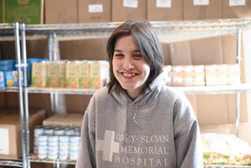 A person wearing a grey sweater smiles as they stand in a food bank.