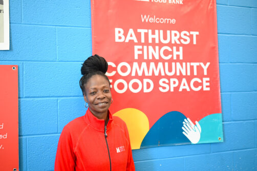 A person in a red North York Harvest Food Bank sweater stands smiling in front of a banner that reads Welcome to Bathurst-Finch Community Food Space