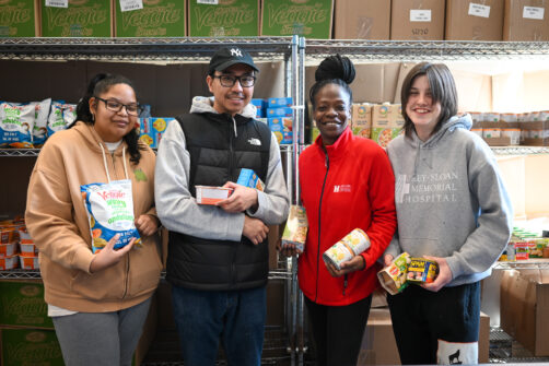 Four food bank staff hold various food items standing in front of the shelves at a food bank.