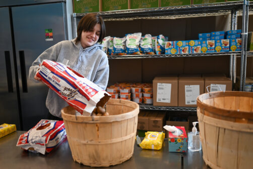 A bag of potatoes are poured into a wooden bin with shelves of food visible in the background.