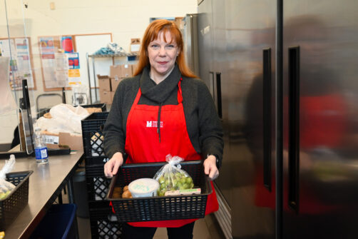A volunteer wearing a North York Harvest Food Bank apron holds a crate of fresh food in a food bank setting.