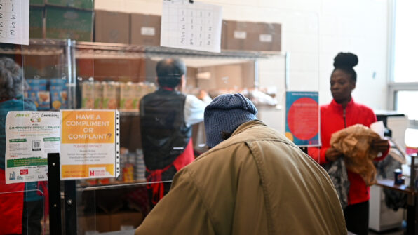 An older adult is visible from behind as they wait to be served at the food bank. In the background, food bank staff pick food off of the shelves.