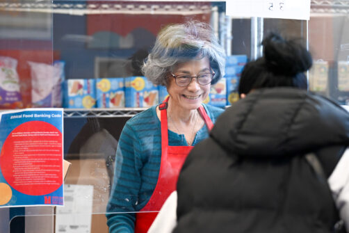 A person stands behind the counter smiling at a food bank as they serve a client.