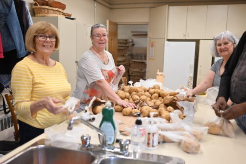 Three women stand around a counter full of potatoes, sorting them for distribution at a food bank.