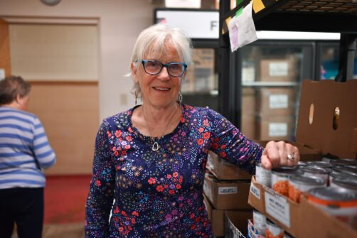 A woman with glasses rests her hand on a stack of non-perishable canned beans in a food bank setting.