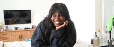 A woman with long hair leans on a kitchen counter, smiling.