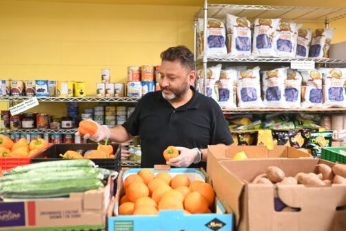 A man inspects oranges for distribution at a food bank, with shelves on non-perishables behind him.