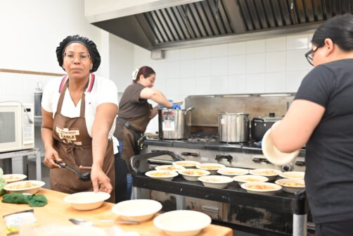 A woman wearing a brown St. Philip Neri's Table apron places a bowl on the counter in a kitchen, ready to be filled with homemade soup. In the background, volunteers load soup onto trays and stir a pot.