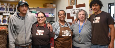 Five volunteers with St. Philip Neri's Table stand inside the food bank with their arms around each other's shoulders, smiling.