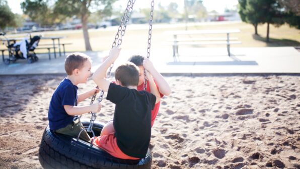 Three children play on a tire swing in a sandy playground setting.