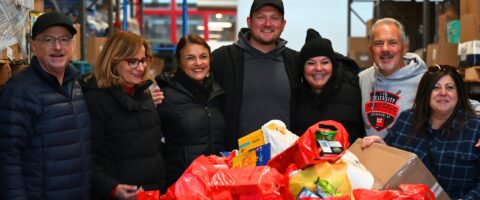 Seven people stand around a large container of donated food at a food bank warehouse, smiling and linking arms.