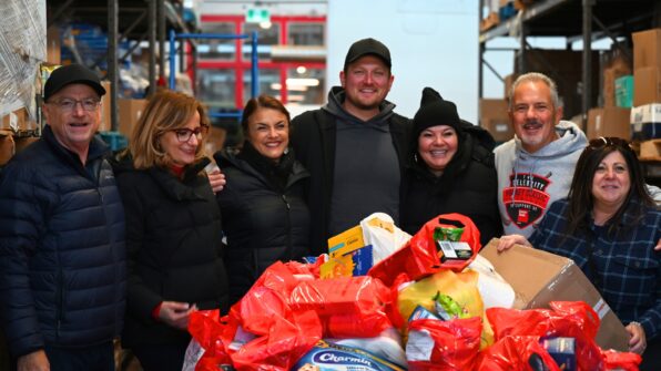 Seven people stand around a large container of donated food at a food bank warehouse, smiling and linking arms.