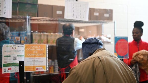 An older woman wearing a hat and winter jacket can be seen from behind as she waits for food to be served at a food bank.