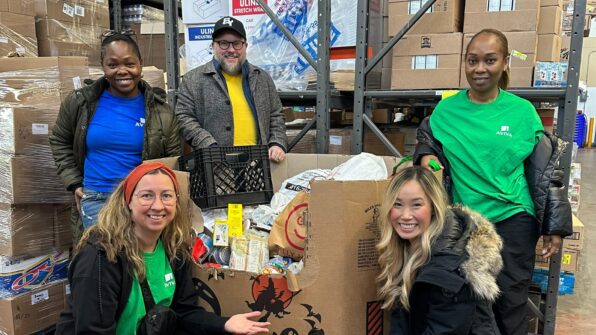 Five people pose for a photo around a large box of donated food items inside of a food bank warehouse.