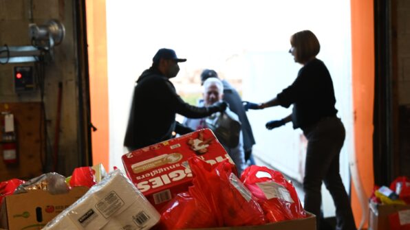 In the foreground is a box of donated items, in the background a person can be seen passing more items for donation in a grocery bag to a food bank employee.
