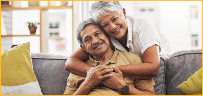An older man sits on the couch with an older woman embracing him from behind. They are both smiling.