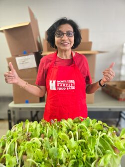 A person wearing a red North York Harvest Food Bank apron holds their thumbs up and smiles with fresh lettuce visible in front of her.