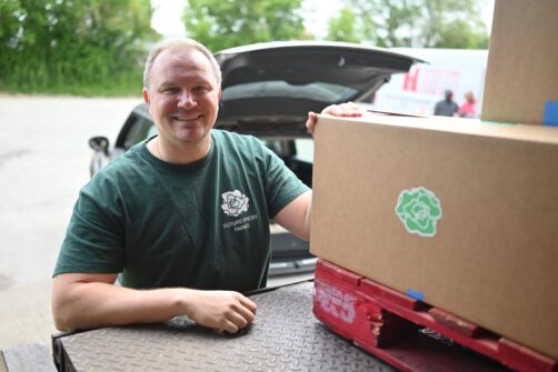 A man wearing a green branded shirt stands at the back of the warehouse next to boxes of food being delivered.