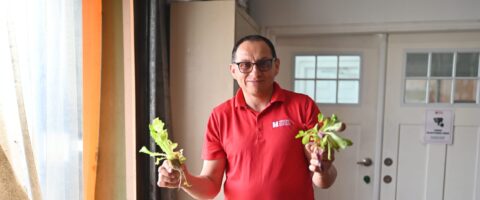 A person wearing a red North York Harvest shirt holds lettuce in a warehouse setting with boxes beside him.