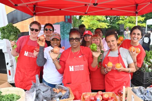 Nine people stand behind a booth at an outdoor market holding fruits and vegetables. Some are wearing North York Harvest Food Bank aprons.