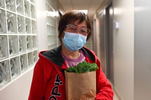 A person wearing a mask holds a bag of fresh lettuce, smiling.