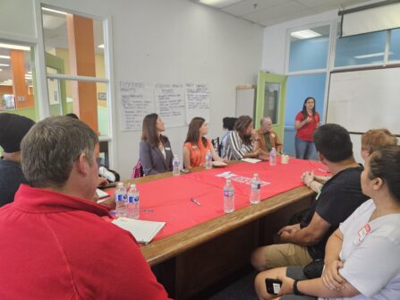 A group of people sit around a table at North York Harvest Food Bank while a person at the front of the room leads a discussion and writes on a whiteboard.