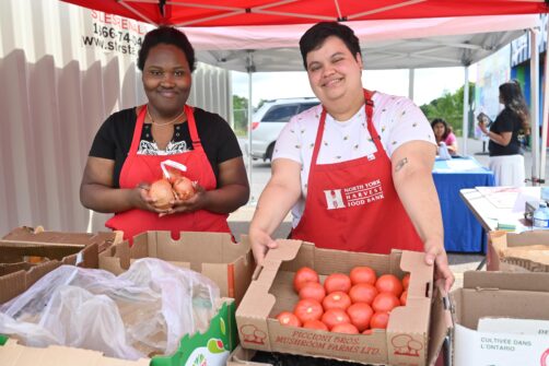 Two people wearing red North York Harvest Food Bank aprons stand at an outdoor market in front of trays of produce.