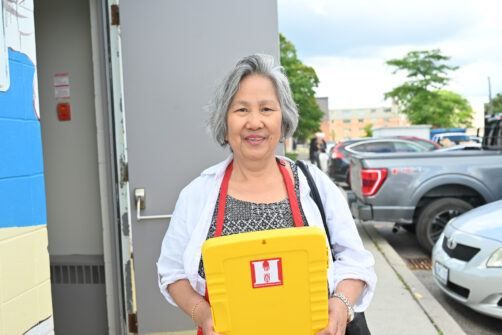 A woman holding a yellow clipboard with a red North York Harvest Food Bank sticker on it stands outside.