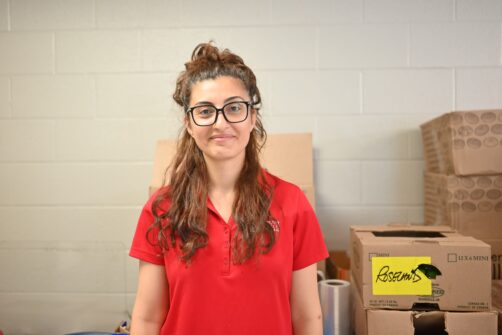 A woman with long brown hair and glasses wearing a red North York Harvest Food Bank shirt smiles.