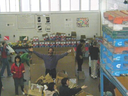 A man stands in the middle of a busy North York Harvest Food Bank warehouse with his arms outstretched.