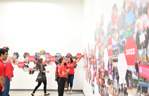 A group of people stand in front of a wall with a timeline in the shape of interconnected honeycombs, weaving together pictures and text. 