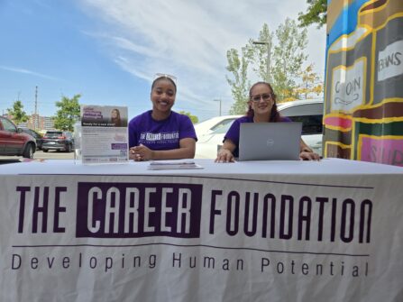 Two people sit at an information table outside, the tablecloth reads The Career Foundation.