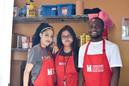 Three people wearing red North York Harvest Food Bank aprons smile in a food bank setting.