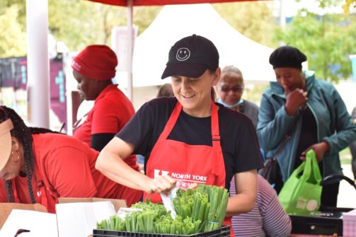 A person wearing a hat with a smiley face on it, and a North York Harvest Food Bank apron serves green onions to a customer at an outdoor market.