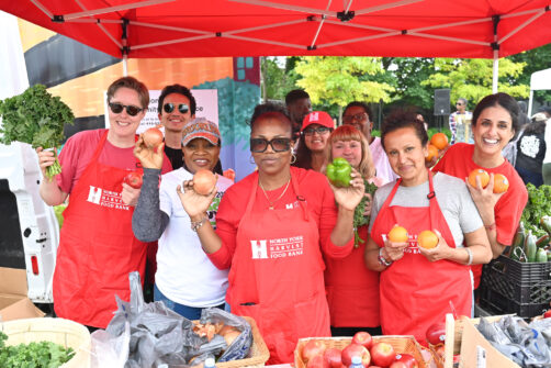 A group of people wearing red North York Harvest Food Bank aprons hold various fruits and vegetables at an outdoor market.