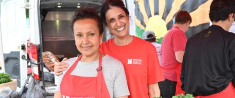 Two people wearing red North York Harvest Food Bank clothing smile at an outdoor market, with fresh produce visible in the foreground.
